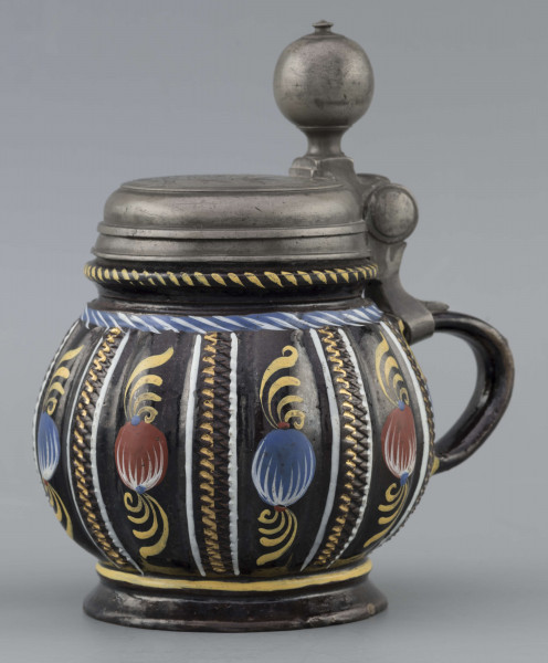 S/CS/595/ML - Kufel  kulisty, o niskiej konicznej stopie i niskiej szyi z pierścieniem, ucho taśmowe w kształcie „C”, pokrywka cynowa. Korpus pionowo kanelowany, podzielony na pasy na przemian gładkie i rytowane w ukośną kratkę. Pokryty szkliwem w kolorze ciemnego kobaltu, zdobiony malowanymi ornamentami podkreślającymi podziały powierzchni, w kolorach: czerwień, żółcień, błękit, biel. Pokrywka płaska, z pierścieniami i uchwytem kulkowym, na zawiasie umocowanym do ucha. Na wierzchu pokrywki grawerunek przedstawiający zawiły ornament – być może monogram spleciony z liter JGL – pod koroną, w uproszczonym wieńcu, z datą 1716.  