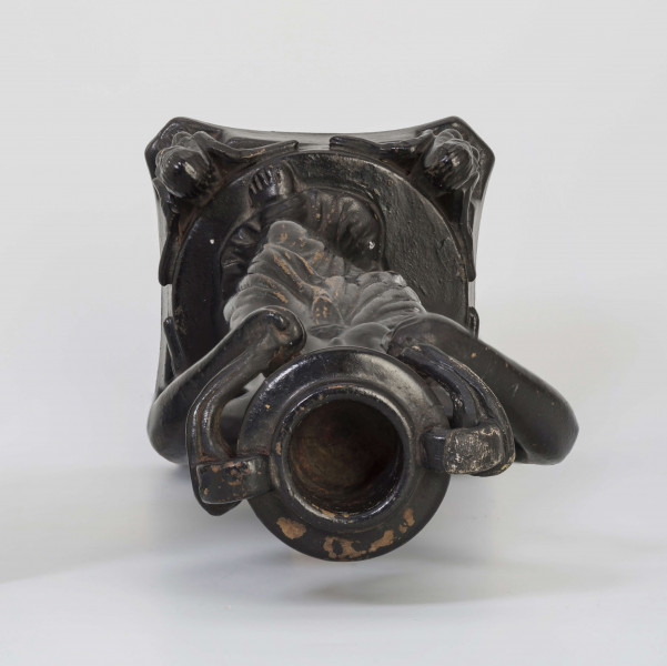 Świecznik figuralny z kariatydą; cokół czworoboczny górą przechodzący w koło, w narożach figurki sfinksów, na nim stojąca postać kobiety z wazonem na głowie, który podtrzymuje uniesionymi rękami. Kobieta do pasa naga, ubrana jest w fałdzistą szatę przwiązaną u góry, na głowie chusta opadajaca na ramiona. Całość malowana lakierem na czarno.
