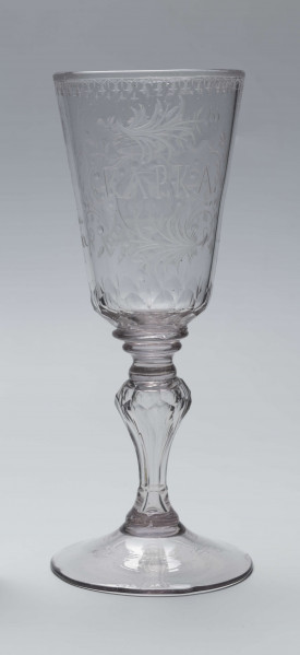 S/CS/244/ML - Stopa okrągła, lekko uniesiona, trzon tralkowy, z pęcherzykiem powietrza, fasetowany, od czaszy oddzielony pierścieniem i dwoma talerzykami, czasza stozkowata, u nasady zaokrąglona, szlifowana w dwa pionowe rzędy owalnych faset. Dekoracja rytowana: na czaszy dwustronna - z jednej strony owalna tarcza z parą gołąbków połączonych dziobkami stojących na serduszkach w rozbudowanym kartuszu z uszakami, z którego wyrastaja gałązki; po drugiej stronie litery 