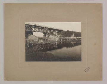 ML/H/F/26/4 ML/H/F/26/4 - Fotografia przedstawia drewniany most na rzece, widoczne zasieki wykonane z drzewa i drutu kolczastego. Na skarpie po drugiej stronie rzek i widoczne sylwetki mężczyzn. Fotografia naklejona na szarą tekturę z wytłoczoną ramką. W prawym dolnym rogu pieczęć zakładu fotograficznego S. Zawadzkiego z Lublina.