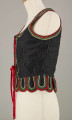 E/16876/ML - Gorset zszyty z 6 kawałków czarnej satyny, podszyty lnianą podszewką. Z przodu rozcięcie usztywnione fiszbinami oraz haftkami do przewlekania wstążeczki. Od pasa dookoła opadają klapki „taszki”. Przód wzdłuż rozcięcie wycięcie wokół szyi, brzegi pach i taszki ozdobione czerwoną, zieloną i białą wełnianą krepinką. Plecy zdobione ornamentem wykonanym ściegiem krzyżykowym. Szyty ręcznie.