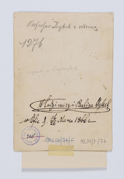 ML/H/F/74 - Fotografia przedstawia rodziców, a pomiędzy nimi stojącą trójkę dzieci. Kobieta w ciemnej sukni okryta szalem, na głowie ma płaski toczek z futra karakułów. Obok na krześle siedzi mężczyzna z dość długimi, gładko zaczesanymi włosami, założonymi za uszy. Ma długą brodę i wąsy. Ubrany w w ciemny garnitur i długi, ciemny płaszcz. Dziewczynka trzyma w ręku lalkę. 
Na odwrociu napis: 