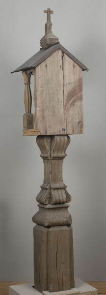 E/8062/1/ML - Kapliczka słupowa wykonana ręcznie siekierą i dłutami. Dębowy, profilowany ozdobnie słup w formie czterograniastej kolumny. Baza przy podstawie spróchniała, ma wgłębione narożniki. Żłobienia, głębokości 3,5 cm, uwypuklają płaszczyzny. Na przedniej ścianie wyryty napis: „Obrobił i postawił własnemi rencznie Jan Oliński dnia 17 marca 1933 r.”. Baza zakończona elementem o przekroju odwróconego trapezu wys. 17 cm i podstawie 26 cm, o zaokrąglonych dolnych narożnikach. Na nim trzon ozdobny czworograniasty, wys. 18 cm. Podstawa trzonu i głowica 4 esowatych żeberek, szerokości 12 cm. Na bocznych pełnych ściankach żeberek uproszony liść akantu. Na głowicach – kapliczka ażurowa. Kolumienki i szczyt brzozowe. Baza kolumny ma kształt graniastosłupa (wys. 12 cm, szer. 7 cm); trzon walcowaty (wys. 22 cm, szer. 8 cm), wybrzuszony, z pionowymi szerokimi rowkowaniami. Głowica (wys. 17 cm), utworzona z uproszczonych liści akantu i dwu złączonych w podstawy rozbieżnych wolut. Szczyt – tympanon ze śladami błękitnej polichromii, ozdobiony perliście małymi sześcianami o zaokrąglonych dolnych krawędziach. U szczytu drewniana kotara (lambrekin) z udrapowanych łukowatych żłobień, w środku ujęty prostą taśmą. Dach dwuspadowy, sosnowy o podciętych i podwiniętych krawędziach dolnych, pokryty blachą. Plecy i podstawa kapliczki – proste, sosnowe.