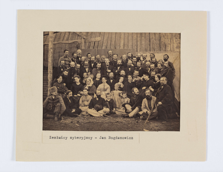 ML/H/561/1 - Fotografia grupowa zesłańców syberyjskich. Widoczne sylwetki dorosłych mężczyzn i kobiet oraz dzieci. Wszyscy ubrani w stroje z epoki. Pozujący do zdjęcia ustawieni w kilka rzędów. Osoby na pierwszym planie siedzą na rozłożnym na ziemi kocu. W tle ściana drewnianego budynku. Spośród 57 postaci rozpoznanych 40 osób.