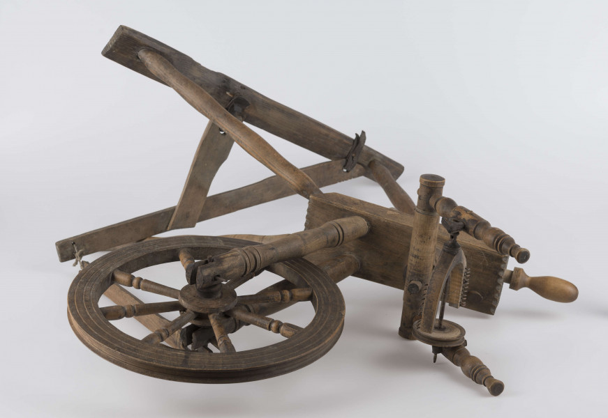 E/16647/ML - Kołowrotek poziomy, drewniany, ozdobnie toczony. Drewno niemalowane. Podstawa to 3 skośnie ustawione nóżki. Dwie z nich osadzone w wąskiej listwie. Górne końce nóżek osadzone w prostokątnej pochyłej grubej desce (11 x 32 x 4 cm). Stanowi ona podstawę do osadzenia koła napędowego i szpuli zwijającej nici. W niej zamocowany uchwyt połączony ze śrubą, która reguluje położenie szpuli. Koło osadzone na metalowej ośce posiada 8 toczonych ozdobnie szprych. Jeden koniec ośki połączony pionową ruchomą listewką z pedałem wprawiającym w ruch kołowrotek. Szpula osadzona również na metalowej ośce.