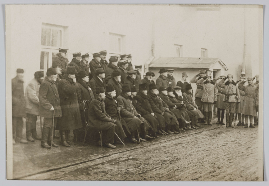 ML/H/310 - Na fotografii grupa weteranów powstania styczniowego, licząca 34 osoby. Ustawieni są w trzech rządach: pierwszy rząd siedzi na krzesłach, drugi stoi, trzeci rząd stoi na podwyższeniu. Część mężczyzn ubrana w czamaraki. Z prawej strony salutujące wojsko. Fotografia wykonana na tle budynków. Na odwrociu wykropkowane linie jak na pocztówkach oraz odręczny napis: Powstańcy z 1863 r.