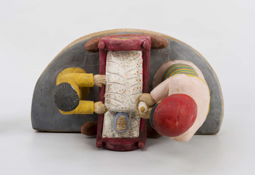 Rzeźba w drewnie lipowym, polichromowana, przedstawiająca postacie matki i dziecka przy kołysce z niemowlęciem. Całość zakomponowana na postumencie wys. 4,4 cm i dł. 22 cm, o kształcie z przodu półokrągłym. Pośrodku usytuowana kołyska na biegunach, wys. 9 cm i dł. 11,2 cm. Kołyska czerwona, ścianki szczytowe ozdobione bukietami składającymi się z trzech wielopłatkowych kwiatów w kolorze żółtym i tulipanowych zielonych liśćmi. W kołysce, w beciku białym, w czerwono-zielone kropki, dziecko, w niebieskiej chustce na głowie. Z lewej strony kołyski postać matki, w długiej jasnoróżowej sukni, w pasie przepasanej fartuchem w żółto-zielono-pomarańczowo-niebieskie poziome pasy. Twarz płaska, nos długi prosty, usta wąskie małe, oczy owalne o podkreślonych białych białkach i czarnych źrenicach, brwi łukowate czarne. Na głowie czerwona chustka zawiązana pod brodą. Spod niej wystają czarne włosy. W rękach trzyma butelkę z mlekiem. Z prawej strony kołyski stoi dziecko w żółtej sukience z niebieskim kołnierzykiem. Twarz dziecka płaska, nos prosty wąski, usta małe, oczy owalne, brwi proste czarne. Włosy czarne, proste sięgające do ramion, zaczesane z grzywką na czole. Ręce wsparte na kołysce. 