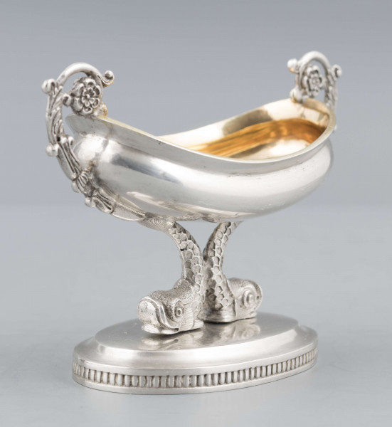 S/Mt/511/ML - Solniczka w formie łódki wspartej na dwóch delfinach, z owalną podstawą, z dwoma uchami z wici roślinnej. Brzeg czarki wywinięty, podstawa lekko podniesiona a jej brzeg zdobiony pionowym karbowaniem. Na spodzie sygnatura:
- imienna 