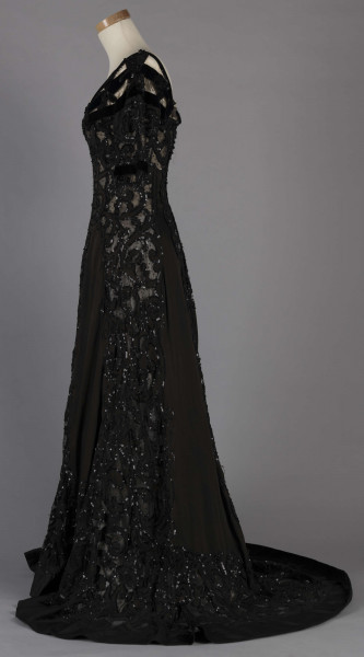 S/T/243/ML - Suknia wizytowa, czarna, uszyta z atłasu jedwabnego, jedwabnego tiulu i aksamitu, haftowana cekinami. Krótki rękaw z jedwabnego czarnego aksamitu, dekolt prostokatny, dość głęboki, wykończony wstawką haftowaną i aksamitem. Kolory są kontrastowe. Spodnia warstwa sukni, z białej tafty i muślinu, widoczna w partiach sukni szytych z czarnego tiulu, dodaje całości elegancji. Brzegi dekoracyjnej wici z tafty zaakcentowane naszytymi cekinami. Suknia do ziemi z tyłu krótki tren. Brak halki. 
