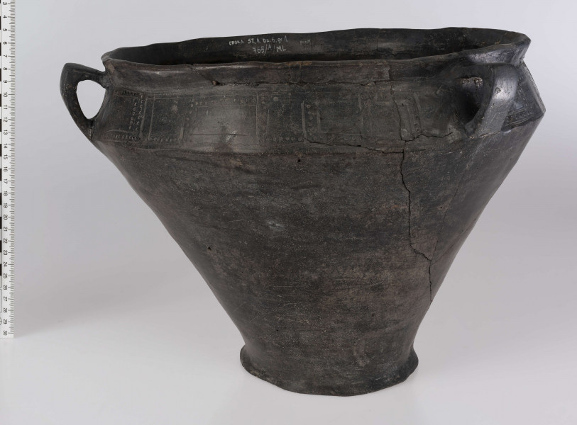 765/A/ML/1 - Waza wyraźnie profilowana, z prostym, odgietym na zewnatrz brzegiem, ostro zaznaczonym załomem brzuśca i wyodrębnionym dnem. Ponad załomem brzuśca występuje ornament w postaci: jodełek, swastyk, tworzonych przez dwie linie ryte i rząd dołków pomiędzy nimi oraz ornament blankowy. Naczynie ma trzy taśmowate ucha kolankowato zgięte. 
