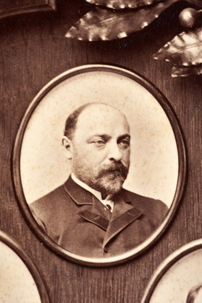 Pośrodku prostokątnego pola o zaokrąglonych narożnikach większa fotografia przyozdobiona wstęgą i gałązkami wawrzynu. Dookoła szesnaście symetrycznie rozmieszczonych mniejszych fotografii. W prawym górnym rogu dr Wacław Lasocki, naczelny lekarz kolei nadwiślańskiej w latach 1875 - 78. W polu ramy w górnej części medalion z płaskorzeźbionym popiersiem z profilu. Przedstawia Leopolda Kronenberga - ojca. Po bokach dwia półleżące aniołki trzymają wieniec laurowy. Po lewej stronie ramy postać robotnika Poleszuka, po prawej pracownika warsztatów kolejowych. W dolnej części front lokomotywy i złoty żeton uprawniający do bezpłatnej jazdy pociągiem I klasy. Fotografia naklejona na tekturę, w dolnej części u dołu sucha pieczęć zakładu fotograficznego.