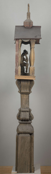 E/8062/1/ML - Kapliczka słupowa wykonana ręcznie siekierą i dłutami. Dębowy, profilowany ozdobnie słup w formie czterograniastej kolumny. Baza przy podstawie spróchniała, ma wgłębione narożniki. Żłobienia, głębokości 3,5 cm, uwypuklają płaszczyzny. Na przedniej ścianie wyryty napis: „Obrobił i postawił własnemi rencznie Jan Oliński dnia 17 marca 1933 r.”. Baza zakończona elementem o przekroju odwróconego trapezu wys. 17 cm i podstawie 26 cm, o zaokrąglonych dolnych narożnikach. Na nim trzon ozdobny czworograniasty, wys. 18 cm. Podstawa trzonu i głowica 4 esowatych żeberek, szerokości 12 cm. Na bocznych pełnych ściankach żeberek uproszony liść akantu. Na głowicach – kapliczka ażurowa. Kolumienki i szczyt brzozowe. Baza kolumny ma kształt graniastosłupa (wys. 12 cm, szer. 7 cm); trzon walcowaty (wys. 22 cm, szer. 8 cm), wybrzuszony, z pionowymi szerokimi rowkowaniami. Głowica (wys. 17 cm), utworzona z uproszczonych liści akantu i dwu złączonych w podstawy rozbieżnych wolut. Szczyt – tympanon ze śladami błękitnej polichromii, ozdobiony perliście małymi sześcianami o zaokrąglonych dolnych krawędziach. U szczytu drewniana kotara (lambrekin) z udrapowanych łukowatych żłobień, w środku ujęty prostą taśmą. Dach dwuspadowy, sosnowy o podciętych i podwiniętych krawędziach dolnych, pokryty blachą. Plecy i podstawa kapliczki – proste, sosnowe.