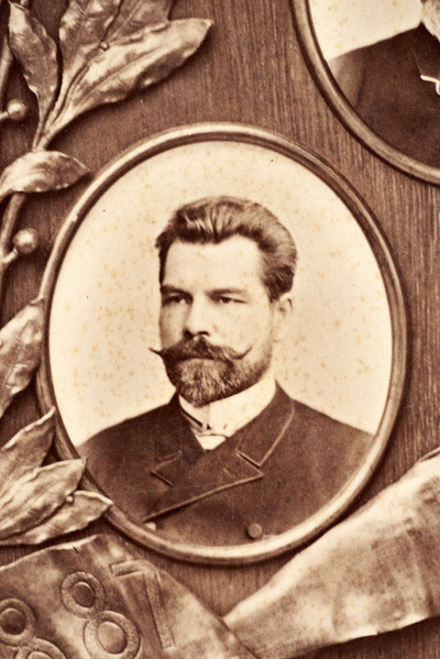 Pośrodku prostokątnego pola o zaokrąglonych narożnikach większa fotografia przyozdobiona wstęgą i gałązkami wawrzynu. Dookoła szesnaście symetrycznie rozmieszczonych mniejszych fotografii. W prawym górnym rogu dr Wacław Lasocki, naczelny lekarz kolei nadwiślańskiej w latach 1875 - 78. W polu ramy w górnej części medalion z płaskorzeźbionym popiersiem z profilu. Przedstawia Leopolda Kronenberga - ojca. Po bokach dwia półleżące aniołki trzymają wieniec laurowy. Po lewej stronie ramy postać robotnika Poleszuka, po prawej pracownika warsztatów kolejowych. W dolnej części front lokomotywy i złoty żeton uprawniający do bezpłatnej jazdy pociągiem I klasy. Fotografia naklejona na tekturę, w dolnej części u dołu sucha pieczęć zakładu fotograficznego.