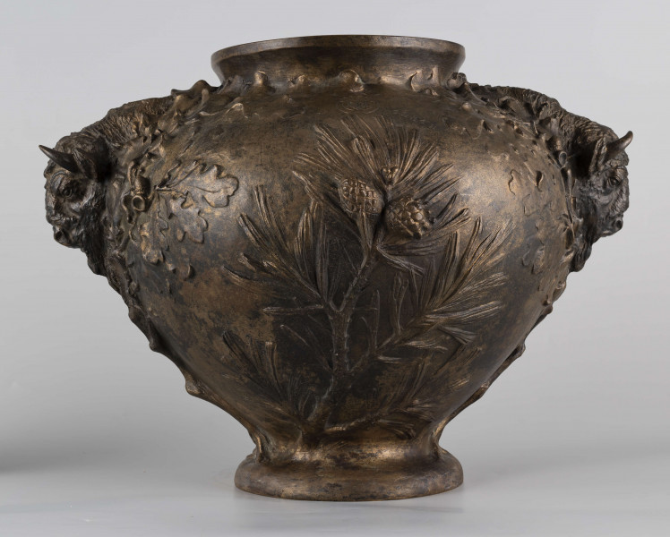 S/Mt/427/ML - Waza w kształcie spłaszczonej kuli, z niską, kolistą stopą i krótką szyją o lekko wywiniętym brzegu. Powierzchnia zdobiona reliefem: po bokach dwa uchwyty w formie głów żubrów, wokół nich gałązki dębu, pośrodku korpusu gałązki sosny z szyszkami wyrastające ze stopy. U góry na brzuścu rytowany napis majuskułą 