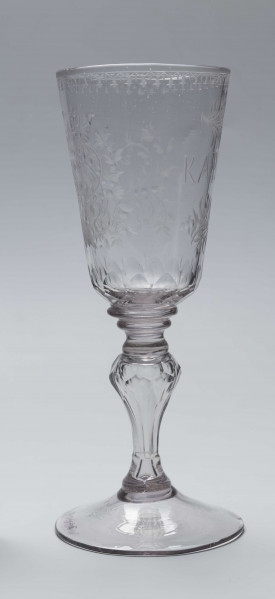 S/CS/244/ML - Stopa okrągła, lekko uniesiona, trzon tralkowy, z pęcherzykiem powietrza, fasetowany, od czaszy oddzielony pierścieniem i dwoma talerzykami, czasza stozkowata, u nasady zaokrąglona, szlifowana w dwa pionowe rzędy owalnych faset. Dekoracja rytowana: na czaszy dwustronna - z jednej strony owalna tarcza z parą gołąbków połączonych dziobkami stojących na serduszkach w rozbudowanym kartuszu z uszakami, z którego wyrastaja gałązki; po drugiej stronie litery 