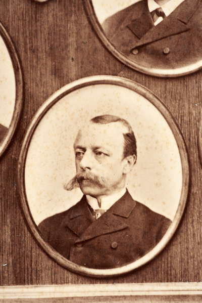 Pośrodku prostokątnego pola o zaokrąglonych narożnikach większa fotografia przyozdobiona wstęgą i gałązkami wawrzynu. Dookoła szesnaście symetrycznie rozmieszczonych mniejszych fotografii. W prawym górnym rogu dr Wacław Lasocki, naczelny lekarz kolei nadwiślańskiej w latach 1875 - 78. W polu ramy w górnej części medalion z płaskorzeźbionym popiersiem z profilu. Przedstawia Leopolda Kronenberga - ojca. Po bokach dwia półleżące aniołki trzymają wieniec laurowy. Po lewej stronie ramy postać robotnika Poleszuka, po prawej pracownika warsztatów kolejowych. W dolnej części front lokomotywy i złoty żeton uprawniający do bezpłatnej jazdy pociągiem I klasy. Fotografia naklejona na tekturę, w dolnej części u dołu sucha pieczęć zakładu fotograficznego.