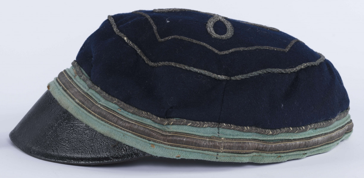 ML/H/2208 - Czapka, czyli tzw. dekiel członka Polskiej Akademickiej Korporacji Astrea działającej na Katolickim Uniwersytecie Lubelskim w latach 1923-1939. Nakrycie głowy w barwach korporanckich, uszyte z nieusztywnianego granatowego sukna, z daszkiem z czarnej skóry, z otokiem w kolorach błękitnym i srebrnym. Na zewnętrznej stronie denka w centralnej części wyszyty fabrycznie srebrną nicią znak korporacji (stylizowany na gwiazdę, w postaci połączonych ze sobą ozdobnych czterech liter „A”) umieszczony wewnątrz haftowanego oktogramu otoczonego kołem z naszytego ozdobnego sznureczka. Po bokach widoczne miejsca zszycia materiału. Wewnątrz podszewka w kolorze granatowym oraz wzmacniający skórzany pasek w kolorze brązowym naokoło.