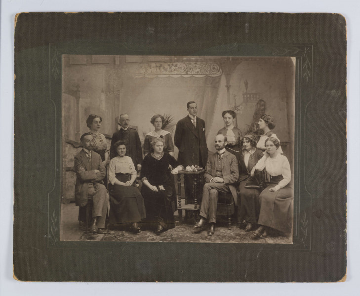 ML/H/F/267 - Fotografia wykonana w atelier na tle panelu z kotarą, na podłodze dywan. Do zdjęcia pozuje 12 osób: 8 kobiet i 4 mężczyzn. Od lewej siedzi Władysław Kunicki, przy stoliku Romana Zapasiewicz, za stolikiem Tołwiński, obok z prawej Józefa Kunicka, z lewej Rennard.
Fotografia naklejona na tekturę w odcieniu szarej zieleni.
Na odwrociu nazwiska niektórych osób wypisane ołówkiem. 