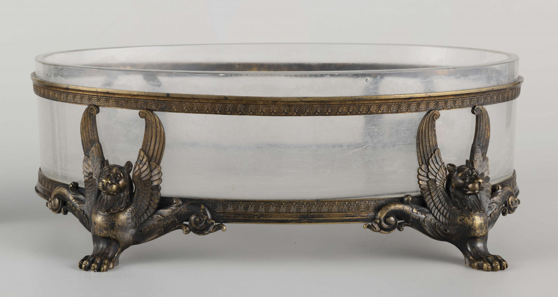 S/CS/1063/ML - Naczynie ze szkła bezbarwnego, grubościennego, owalne, z płaskim dnem, w oprawie metalowej złożonej z dwóch obręczy połączonych plastycznymi figurkami gryfów na nóżkach. Powierzchnia obręczy zdobiona plastycznym fryzem antykizujacych palmet. Oprawa metalowa patynowana. 
 