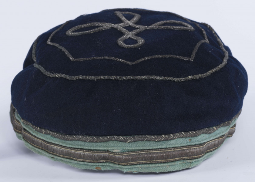 ML/H/2208 - Czapka, czyli tzw. dekiel członka Polskiej Akademickiej Korporacji Astrea działającej na Katolickim Uniwersytecie Lubelskim w latach 1923-1939. Nakrycie głowy w barwach korporanckich, uszyte z nieusztywnianego granatowego sukna, z daszkiem z czarnej skóry, z otokiem w kolorach błękitnym i srebrnym. Na zewnętrznej stronie denka w centralnej części wyszyty fabrycznie srebrną nicią znak korporacji (stylizowany na gwiazdę, w postaci połączonych ze sobą ozdobnych czterech liter „A”) umieszczony wewnątrz haftowanego oktogramu otoczonego kołem z naszytego ozdobnego sznureczka. Po bokach widoczne miejsca zszycia materiału. Wewnątrz podszewka w kolorze granatowym oraz wzmacniający skórzany pasek w kolorze brązowym naokoło.