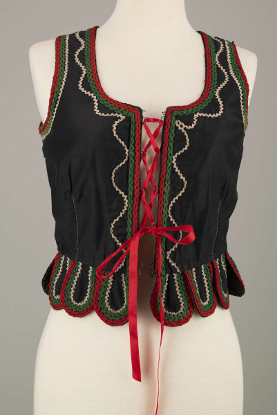 E/16876/ML - Gorset zszyty z 6 kawałków czarnej satyny, podszyty lnianą podszewką. Z przodu rozcięcie usztywnione fiszbinami oraz haftkami do przewlekania wstążeczki. Od pasa dookoła opadają klapki „taszki”. Przód wzdłuż rozcięcie wycięcie wokół szyi, brzegi pach i taszki ozdobione czerwoną, zieloną i białą wełnianą krepinką. Plecy zdobione ornamentem wykonanym ściegiem krzyżykowym. Szyty ręcznie.