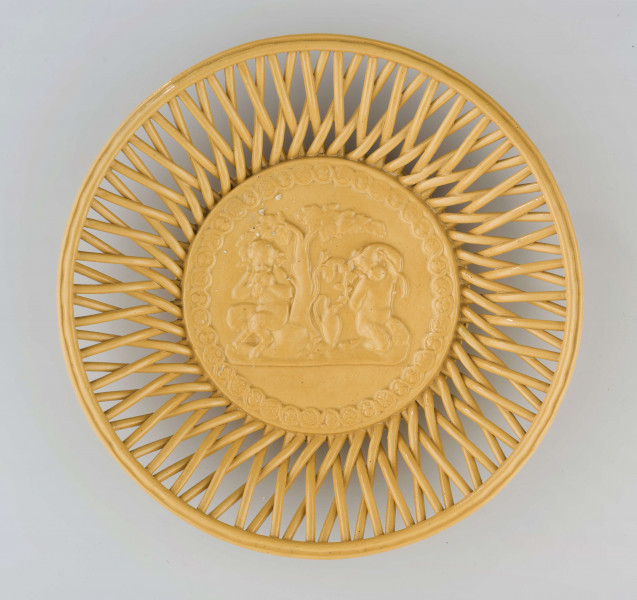  Koszyczek okrągły, płytki; dno płaskie, zdobione plastyczną dekoracją z motywem figuralnym przedstawiającym fauna i amorka pod drzewem; kołnierz ażurowy z okrągłych krzyżujących się wałków połączonych profilowaną obręczą tworzącą krawędź. Całość pokryta połyskliwą glazurą w kolorze ugru.
 