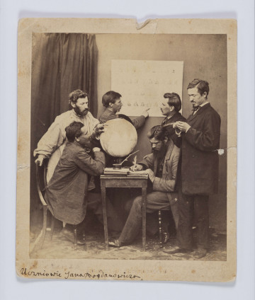 ML/H/F/68 - Jan Bogdanowicz z uczniami na zesłaniu. Fotografia przedstawia sześciu mężczyzn (dwóch siedzących i czterech stojących). Grupa zebrana w pomieszczeniu przy małym kwadratowym stoliku, na którym znajduje się globus, książki, zeszyty i kałamarz z piórem. Jedna z postaci, siedząca po prawej, pisze gęsim piórem. Osoba siedząca naprzeciw wskazuje lewą ręką punkt na globusie. Na ścianie nad stolikiem wisi plansza. Przy niej stoją dwaj mężczyźni - postać po lewej wskazuje coś na planszy prawą dłonią. Nad globusem pochylony jest mężczyzna z brodą w białej długiej koszuli ze stójką (tzw. 