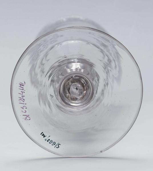 S/CS/244/ML - Stopa okrągła, lekko uniesiona, trzon tralkowy, z pęcherzykiem powietrza, fasetowany, od czaszy oddzielony pierścieniem i dwoma talerzykami, czasza stozkowata, u nasady zaokrąglona, szlifowana w dwa pionowe rzędy owalnych faset. Dekoracja rytowana: na czaszy dwustronna - z jednej strony owalna tarcza z parą gołąbków połączonych dziobkami stojących na serduszkach w rozbudowanym kartuszu z uszakami, z którego wyrastaja gałązki; po drugiej stronie litery 