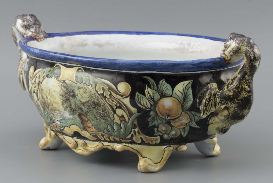 S/CS/90/ML - Żardiniera owalna, na czterech konsolkowych nóżkach połączonych falistymi spływami, wylew z lekko rozchylonym brzegiem, po bokach dwa uchwyty w formie uskrzydlonych smoków, pionowo osadzone. Powierzchni malowana wielobarwnie:  po obu stronach bogate kartusze wolutowo-liściaste z pejzażami  -  jeden z zamkiem obronnym, drugi z wierzbą, po bokach kartuszy festony owoców na ciemno granatowym tle. Wylew obwiedziony błękitnym pasem, wnętrze białobłękitne. 