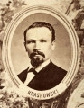 Fotografie w odcieniu sepii umieszczone na tle drzew i roślin, w centralnym miejscu postać kobiety ze sztandarem i aniołkami, wkomponowane widoki budowanej stacji, mostu kolejowego i lokomotywy. Największe zdjęcie w górnej, środkowej części bez podpisu, prawdopodobnie inż. głównej kolei nadwiślańskiej Tadeusz Chrzanowski lub jego następca Walery Titkow. Wśród podobizn pracownicy Biura Głównego Inżyniera: inż. Włodzimierz Stanisławski - pomocnik głównego inżyniera; inż. Feliks Kucharzewski -  pomocnik naczelnika biura; Feliks Kamocki - naczelnik kancelarii; Witold Lanci - naczelnik Wydz. Architektury; dr Wacław Lasocki- służba zdrowia; Władysław Radwan - sprawy zakupu gruntów; inż. Stanisław hr Scipio - projektant mostów kolejowych i jego współpracownicy: inż. Stefan Zieliński, inż. Maurycy Machalski, inż. Feliks Kucharzewski. Fotografia w passe - partout, za szkłem, oprawiona w drewnianą, profilowaną ramę.