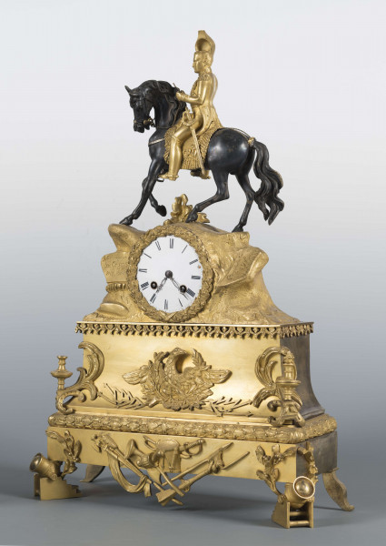 S/Mt/410/ML - Zegar kominkowy z brązu złoconego w stylu empire. Obudowa trzystopniowa, zwężająca się ku górze, na czterech nóżkach w formie armat, złożona z prostokątnej podstawy, części środkowej o wklęsłej linii boków i górnej imitującej skalisty szczyt; podziały podkreślone pasami ornamentu. Powierzchnię zdobią nakładki – w dolnej części panoplia i wieńce laurowe, w środkowej cesarski orzeł, w narożach umieszczono znicze. Pośrodku części górnej wieniec dębowy a w nim umieszczona okrągła tarcza zegarowa pokryta białą emalią, z czarnymi cyframi rzymskimi godzin i oznaczeniami minut. W tarczy dwie ażurowe wskazówki i dwa otwory na osie sprężyn, po bokach tarczy wyryte napisy: 