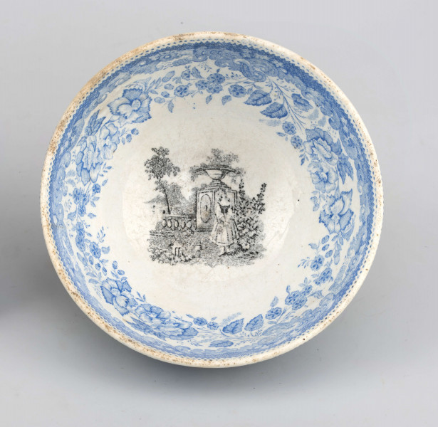 Miseczka półkulista, na niskiej, okrągłej stopce. Zdobiona od strony zewnetrznej i wewnętrznej nadrukiem w kolorze błękitnym z wzorem wolut i ukwieconych gałązek róży i goździków. W lustrze scena w kolorze czarnym przedstawiająca młodą dziewczynę w ogrodzie z elementami 