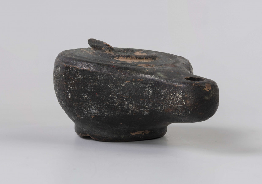 720/A/ML/2 - Lampka oliwna z utłuczonym uszkiem; mały otwór do wlewania oliwy obwiedziony plastycznym wałeczkiem, który jest połączony z uszkiem plastycznym wałeczkiem rozszerzającym się w kierunku uszka; wokół krawędzi dookolny złobek; mały otwór na płomien na wysmukłym, dość mocno wyciągniętym dziubku; dno wyodrębnione, wklęsłe; prawdopodobnie miniatura kultowej, pannońska wzorowana na lampach brązowych