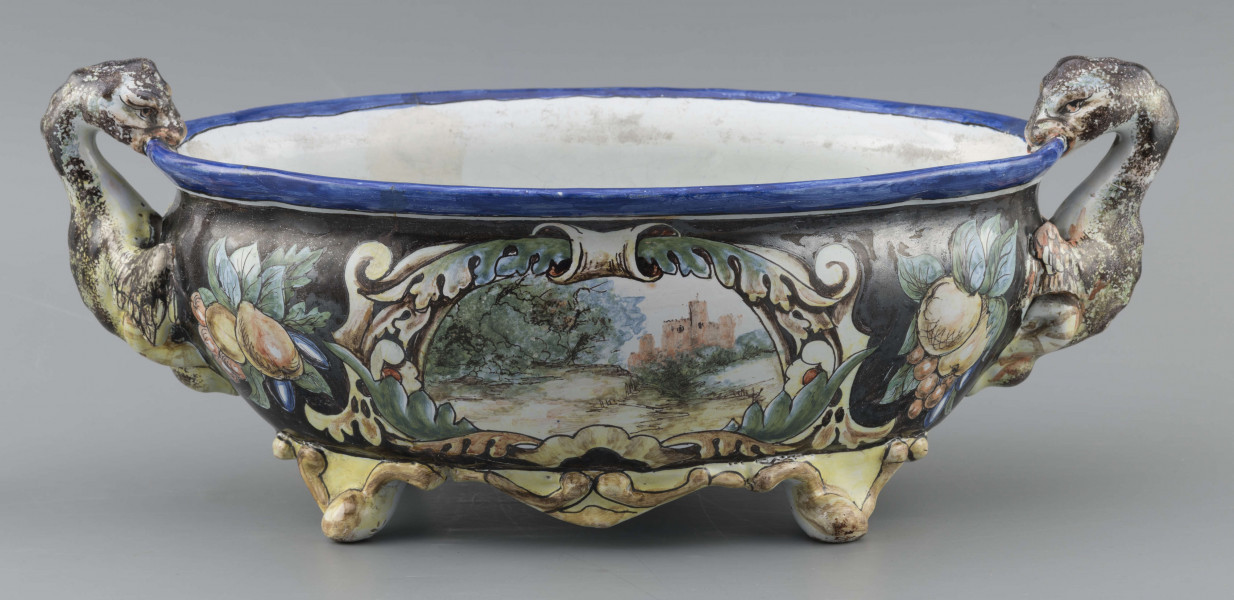 S/CS/90/ML - Żardiniera owalna, na czterech konsolkowych nóżkach połączonych falistymi spływami, wylew z lekko rozchylonym brzegiem, po bokach dwa uchwyty w formie uskrzydlonych smoków, pionowo osadzone. Powierzchni malowana wielobarwnie:  po obu stronach bogate kartusze wolutowo-liściaste z pejzażami  -  jeden z zamkiem obronnym, drugi z wierzbą, po bokach kartuszy festony owoców na ciemno granatowym tle. Wylew obwiedziony błękitnym pasem, wnętrze białobłękitne. 