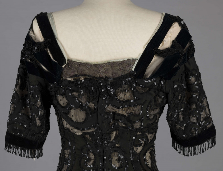 S/T/243/ML - Suknia wizytowa, czarna, uszyta z atłasu jedwabnego, jedwabnego tiulu i aksamitu, haftowana cekinami. Krótki rękaw z jedwabnego czarnego aksamitu, dekolt prostokatny, dość głęboki, wykończony wstawką haftowaną i aksamitem. Kolory są kontrastowe. Spodnia warstwa sukni, z białej tafty i muślinu, widoczna w partiach sukni szytych z czarnego tiulu, dodaje całości elegancji. Brzegi dekoracyjnej wici z tafty zaakcentowane naszytymi cekinami. Suknia do ziemi z tyłu krótki tren. Brak halki. 