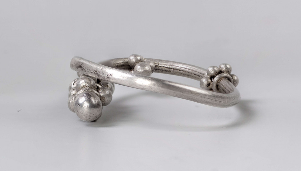 169/A/ML/1 - Zausznica srebrna składa się z kabłąka, na którym zawieszono wisiorek o winogronowatym kształcie. Ozdoby tego typu charakteryzują się kabłąkiem o jednym końcu ucięty prosto, drugim zaś zwiniętym w oczko. W połowie wysokości kabłąka znajdują się wieńce wykonane z granulek ograniczonych pierścieniami z drutu. W dolnej części kabłąka występuje wisiorek uformowany z kilku wieńców granulek zwieńczony granulką osadzoną na pierścieniu z drutu. 