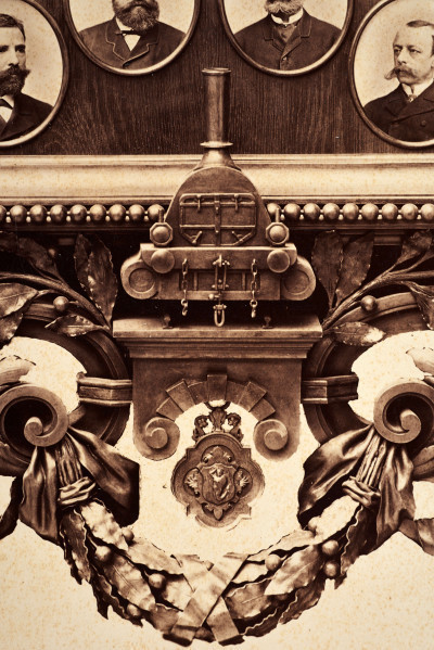 Pośrodku prostokątnego pola o zaokrąglonych narożnikach większa fotografia przyozdobiona wstęgą i gałązkami wawrzynu. Dookoła szesnaście symetrycznie rozmieszczonych mniejszych fotografii. W prawym górnym rogu dr Wacław Lasocki, naczelny lekarz kolei nadwiślańskiej w latach 1875 - 78. W polu ramy w górnej części medalion z płaskorzeźbionym popiersiem z profilu. Przedstawia Leopolda Kronenberga - ojca. Po bokach dwia półleżące aniołki trzymają wieniec laurowy. Po lewej stronie ramy postać robotnika Poleszuka, po prawej pracownika warsztatów kolejowych. W dolnej części front lokomotywy i złoty żeton uprawniający do bezpłatnej jazdy pociągiem I klasy. Fotografia naklejona na tekturę, w dolnej części u dołu sucha pieczęć zakładu fotograficznego.