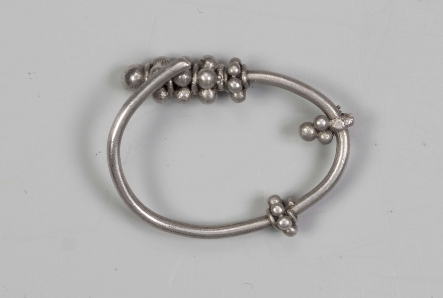 169/A/ML/1 - Zausznica srebrna składa się z kabłąka, na którym zawieszono wisiorek o winogronowatym kształcie. Ozdoby tego typu charakteryzują się kabłąkiem o jednym końcu ucięty prosto, drugim zaś zwiniętym w oczko. W połowie wysokości kabłąka znajdują się wieńce wykonane z granulek ograniczonych pierścieniami z drutu. W dolnej części kabłąka występuje wisiorek uformowany z kilku wieńców granulek zwieńczony granulką osadzoną na pierścieniu z drutu. 