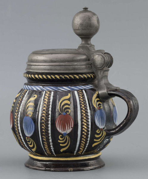 S/CS/595/ML - Kufel  kulisty, o niskiej konicznej stopie i niskiej szyi z pierścieniem, ucho taśmowe w kształcie „C”, pokrywka cynowa. Korpus pionowo kanelowany, podzielony na pasy na przemian gładkie i rytowane w ukośną kratkę. Pokryty szkliwem w kolorze ciemnego kobaltu, zdobiony malowanymi ornamentami podkreślającymi podziały powierzchni, w kolorach: czerwień, żółcień, błękit, biel. Pokrywka płaska, z pierścieniami i uchwytem kulkowym, na zawiasie umocowanym do ucha. Na wierzchu pokrywki grawerunek przedstawiający zawiły ornament – być może monogram spleciony z liter JGL – pod koroną, w uproszczonym wieńcu, z datą 1716.  