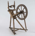 E/16647/ML - Kołowrotek poziomy, drewniany, ozdobnie toczony. Drewno niemalowane. Podstawa to 3 skośnie ustawione nóżki. Dwie z nich osadzone w wąskiej listwie. Górne końce nóżek osadzone w prostokątnej pochyłej grubej desce (11 x 32 x 4 cm). Stanowi ona podstawę do osadzenia koła napędowego i szpuli zwijającej nici. W niej zamocowany uchwyt połączony ze śrubą, która reguluje położenie szpuli. Koło osadzone na metalowej ośce posiada 8 toczonych ozdobnie szprych. Jeden koniec ośki połączony pionową ruchomą listewką z pedałem wprawiającym w ruch kołowrotek. Szpula osadzona również na metalowej ośce.