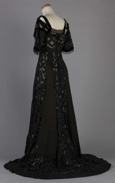 S/T/243/ML - Suknia wizytowa, czarna, uszyta z atłasu jedwabnego, jedwabnego tiulu i aksamitu, haftowana cekinami. Krótki rękaw z jedwabnego czarnego aksamitu, dekolt prostokatny, dość głęboki, wykończony wstawką haftowaną i aksamitem. Kolory są kontrastowe. Spodnia warstwa sukni, z białej tafty i muślinu, widoczna w partiach sukni szytych z czarnego tiulu, dodaje całości elegancji. Brzegi dekoracyjnej wici z tafty zaakcentowane naszytymi cekinami. Suknia do ziemi z tyłu krótki tren. Brak halki. 