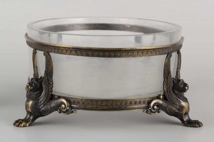 S/CS/1063/ML - Naczynie ze szkła bezbarwnego, grubościennego, owalne, z płaskim dnem, w oprawie metalowej złożonej z dwóch obręczy połączonych plastycznymi figurkami gryfów na nóżkach. Powierzchnia obręczy zdobiona plastycznym fryzem antykizujacych palmet. Oprawa metalowa patynowana. 
 