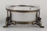 S/CS/1063/ML - Naczynie ze szkła bezbarwnego, grubościennego, owalne, z płaskim dnem, w oprawie metalowej złożonej z dwóch obręczy połączonych plastycznymi figurkami gryfów na nóżkach. Powierzchnia obręczy zdobiona plastycznym fryzem antykizujacych palmet. Oprawa metalowa patynowana. 
 