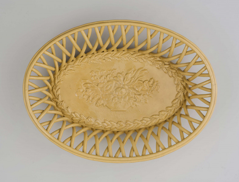 Koszyczek owalny głęboki; dno pełne od zewnątrz obwiedzione warkoczem, boki z okrągłych wałków przeplecionych ukośnie, połączonych profilowaną obręczą tworzącą krawędź. W lustrze (na dnie) dekoracja reliefowa: pośrodku bukiet kwiatów w wazonie, wokół brzegu wić laurowa z owocami. Całość pokryta połyskliwą glazurą w kolorze jasnego ugru.
 