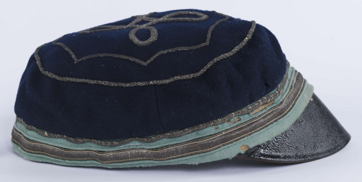 ML/H/2208 - Czapka, czyli tzw. dekiel członka Polskiej Akademickiej Korporacji Astrea działającej na Katolickim Uniwersytecie Lubelskim w latach 1923-1939. Nakrycie głowy w barwach korporanckich, uszyte z nieusztywnianego granatowego sukna, z daszkiem z czarnej skóry, z otokiem w kolorach błękitnym i srebrnym. Na zewnętrznej stronie denka w centralnej części wyszyty fabrycznie srebrną nicią znak korporacji (stylizowany na gwiazdę, w postaci połączonych ze sobą ozdobnych czterech liter „A”) umieszczony wewnątrz haftowanego oktogramu otoczonego kołem z naszytego ozdobnego sznureczka. Po bokach widoczne miejsca zszycia materiału. Wewnątrz podszewka w kolorze granatowym oraz wzmacniający skórzany pasek w kolorze brązowym naokoło.