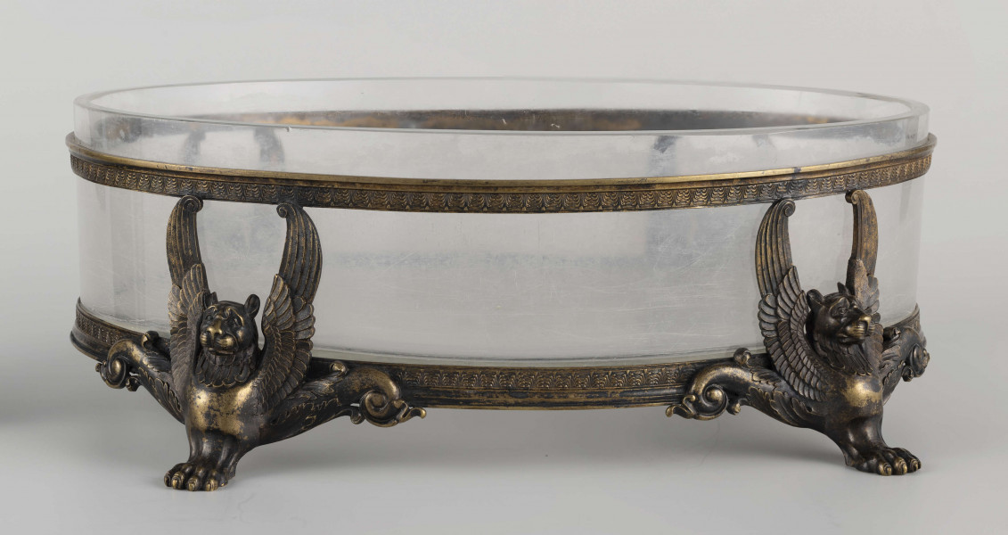 S/CS/1063/ML - Naczynie ze szkła bezbarwnego, grubościennego, owalne, z płaskim dnem, w oprawie metalowej złożonej z dwóch obręczy połączonych plastycznymi figurkami gryfów na nóżkach. Powierzchnia obręczy zdobiona plastycznym fryzem antykizujacych palmet. Oprawa metalowa patynowana. 
 