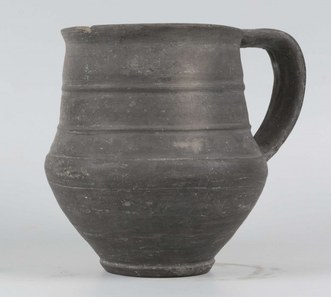 Dno płaskie, wyraźnie wyodrębnione stopką skośnie ściętą ku dołowi; dno wyodrębnione od części przydennej dookolnym wgłębieniem; lekko rozszerzające się ku górze, brzusiec łagodnie zaokraglony, ku górze zwężający się, przechodzący w wysoką, cylindryczną szyjkę; wylew płasko ścięty, rozchylony na zewnątrz; na największej wydętości brzuśca dookolny żłobek; w miejscu przejścia brzuśca w szyję i w połowie szyi dookolny wypukłe listwy; ucho z taśmy środkiem wklęsłej, biegnące od wylewu do załomu brzuśca, szyja zdobiona różnym ornamentem (trzy pasy) przez wygładzenie; powierzchnia gładzona, wyświecona
wymiary dodatkowe: 17,0 cm (obwód); 8,0 cm (śr. dna); 0,3 cm (wys. stopki); 0,5 cm (wys. szyi)