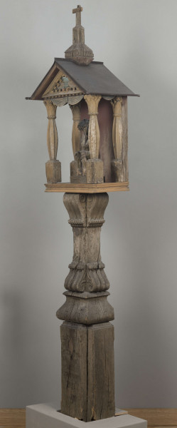 E/8062/1/ML - Kapliczka słupowa wykonana ręcznie siekierą i dłutami. Dębowy, profilowany ozdobnie słup w formie czterograniastej kolumny. Baza przy podstawie spróchniała, ma wgłębione narożniki. Żłobienia, głębokości 3,5 cm, uwypuklają płaszczyzny. Na przedniej ścianie wyryty napis: „Obrobił i postawił własnemi rencznie Jan Oliński dnia 17 marca 1933 r.”. Baza zakończona elementem o przekroju odwróconego trapezu wys. 17 cm i podstawie 26 cm, o zaokrąglonych dolnych narożnikach. Na nim trzon ozdobny czworograniasty, wys. 18 cm. Podstawa trzonu i głowica 4 esowatych żeberek, szerokości 12 cm. Na bocznych pełnych ściankach żeberek uproszony liść akantu. Na głowicach – kapliczka ażurowa. Kolumienki i szczyt brzozowe. Baza kolumny ma kształt graniastosłupa (wys. 12 cm, szer. 7 cm); trzon walcowaty (wys. 22 cm, szer. 8 cm), wybrzuszony, z pionowymi szerokimi rowkowaniami. Głowica (wys. 17 cm), utworzona z uproszczonych liści akantu i dwu złączonych w podstawy rozbieżnych wolut. Szczyt – tympanon ze śladami błękitnej polichromii, ozdobiony perliście małymi sześcianami o zaokrąglonych dolnych krawędziach. U szczytu drewniana kotara (lambrekin) z udrapowanych łukowatych żłobień, w środku ujęty prostą taśmą. Dach dwuspadowy, sosnowy o podciętych i podwiniętych krawędziach dolnych, pokryty blachą. Plecy i podstawa kapliczki – proste, sosnowe.