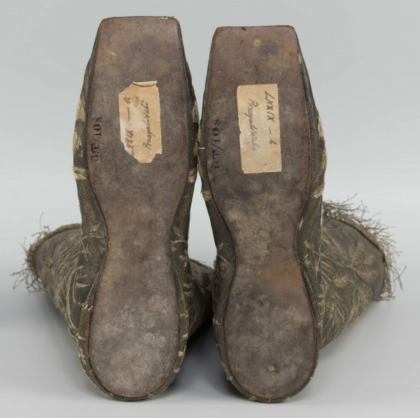 S/T/42/ML - Para butów z wysokimi cholewkami. Cholewka z miękkiej,zielonej skóry,  z dekoracyjnym haftem złotą nicią. Haft z dużym motywem kwiatowym: piwonie, kampanule i drobne kwiatki niezapominajki, tulipany(?), wydłużone liście i wici roślinne, wygięte łukowato.  Wewnatrz buty mają wykończenie białym, cienkim płótnem. Brzegi cholewki obszyte złota koronką z frędzlą. Podeszwy cieńkie z jednego kawałka skóry, bez obcasów, płaskie, z przodu prosto ścięte. Obydwie, skórzane podeszwy mają taki sam wykrój, bez rozróżnienia na lewą i prawą.