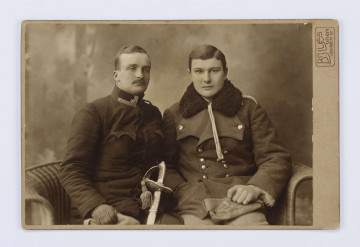 Fotografia Witolda Żyborskiego i Jerzego Pola. Przedstawia dwóch mężczyzn siedzących na wiklinowej kanapie. Mężczyźni ubrani w mundury, ze wzrokiem skierowanym prosto, ukazani en face do kolan. Po lewej stronie Jerzy z krótkimi włosami i wąsami, ze wzrokiem utkwionym w przestrzeń. Trzyma szablę z ozdobną rękojeścią. Z prawej strony Witold Żyborski, z ciemnymi, zaczesanymi na bok włosami. Ubrany w dwurzędową kurtkę wojskową z futrzanym kołnierzem. W lewej ręce trzyma na kolanach czapkę rogatywkę. Fotografia naklejona na sztywną tekturkę. W rogu napis drukowany: B.JJLS/Wien/GRABEN 17. Na odwrocie odręczny napis niebieskim długopisem: Tatuś i Tolo/ Jerzy Pol i wujek Tolo Żyborski.